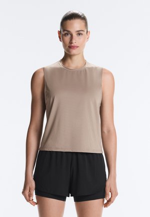 Kvinde med samlet hår bærer beige ærmeløs top og sorte træningsshorts og står mod en ensartet lys baggrund.