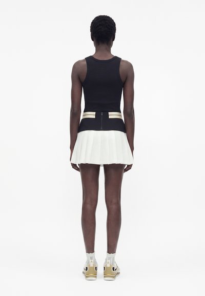 Άτομο με πλάτη, φοράει μαύρο αμάνικο τοπ με μπεζ ρίγες, λευκή πιετέ φούστα, λευκές κάλτσες και χρυσά αθλητικά παπούτσια σε λευκό φόντο.