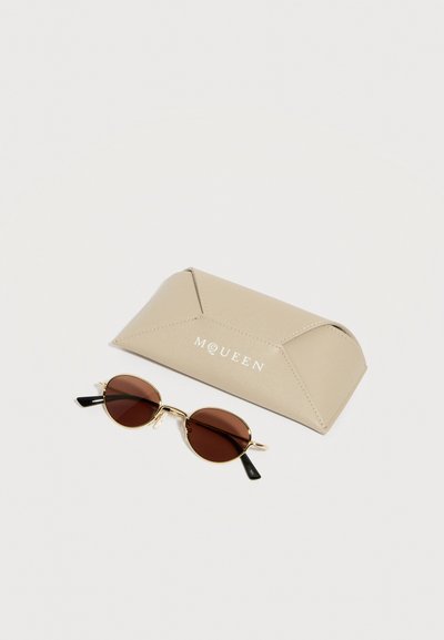 Στρογγυλά γυαλιά ηλίου με χρυσή κορνίζα και καφέ φακούς δίπλα σε θήκη γυαλιών ηλίου McQueen μπεζ χρώματος, σε λευκό φόντο.