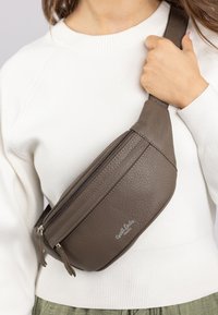 Braune Lederbumstasche mit strukturierter Oberfläche, zwei mit Reißverschluss versehenen Fächern und verstellbarem Gurt, quer über einem weißen Pullover getragen.