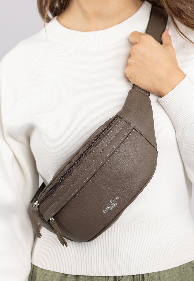 Braune Lederbumstasche mit strukturierter Oberfläche, zwei mit Reißverschluss versehenen Fächern und verstellbarem Gurt, quer über einem weißen Pullover getragen.