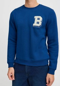 Plava dukserica s okruglim izrezom, s rebrastim manžetama i donjim rubom. Ima veliki, bijeli vezeni znak "B" na prsima. Glatka tekstura.