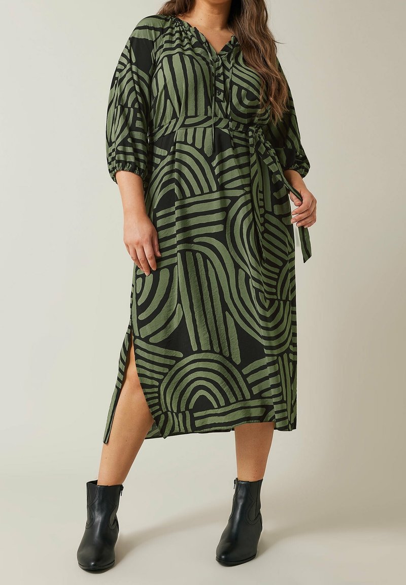Femme portant une robe midi verte et noire à motifs, manches aux coudes et fente latérale, associée à des bottines noires.