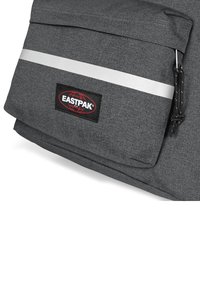 Tumma harmaa reppu, jossa on teksturoitu kangas, suuri läppä, etutasku ja heijastava hopea raita. sisältää Eastpak-logopatchin.