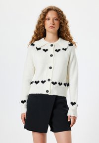 Cárdigan de punto blanco con un patrón de corazones en negro. Presenta un amplio cuello, cinco botones negros y puños y dobladillo acanalados. Combinado con pantalones cortos negros.