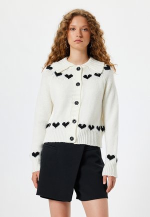 Cárdigan de punto blanco con un patrón de corazones en negro. Presenta un amplio cuello, cinco botones negros y puños y dobladillo acanalados. Combinado con pantalones cortos negros.