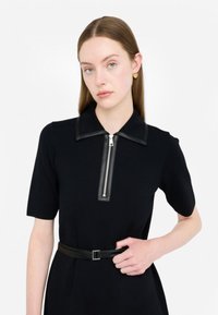 Abito nero con maniche corte, caratterizzato da una cerniera con rifiniture in pelle e una cintura coordinata. Tessuto dalla texture liscia, design del colletto strutturato.