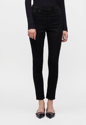 Persona che indossa pantaloni neri slim fit, top nero a maniche lunghe e scarpe nere con punta appuntita, in piedi su sfondo bianco.