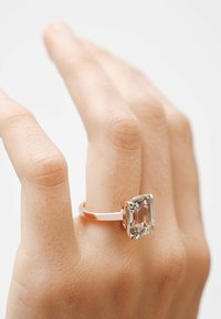 Anillo de oro rosado con una piedra clara, rectangular y facetada, engastada en un ajuste de garras. Banda lisa con un acabado pulido.