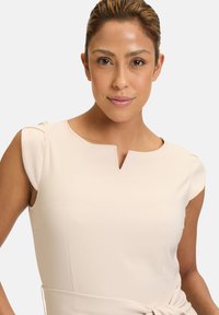 Top beige con scollatura a V, maniche corte e design aderente. Texture liscia con sottili pieghe sulle spalle e senza motivi o hardware visibili.