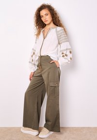 Lässige weiße Bluse mit bestickten Ärmeln, kombiniert mit olivegrünen, weiten Cargo-Hosen. Beiges Sneakers runden das Outfit ab.