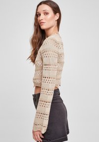 Strikket, kort genser i lys beige med åpne vevmønstre og lange ermer. Kombinert med et svart miniskjørt. Teksturert overflate.