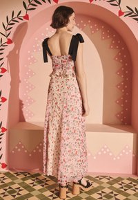 Kvinna som står i en blommig rosa klänning med svarta rosettband och träplattformsklackar mot en rosa bakgrund med hjärtmönstrad båge.