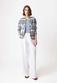 Adīta bumbas jaka zila krāsā ar sarežģītiem rakstiem, apvienota ar gaiši zilu topu un vaļīgām baltām linu biksēm. Sandales ar gludu zoli.