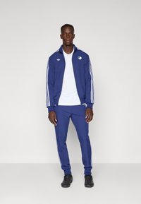 Μπλε αθλ�ητικό φόρμα με λευκές ρίγες, που περιλαμβάνει μπουφάν με φερμουάρ και εφαρμοστά παντελόνια. Φοριέται πάνω από λευκό T-shirt, σετ με μαύρα παπούτσια.