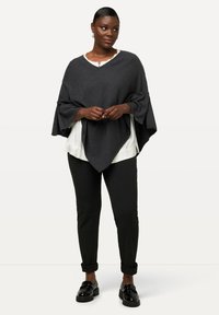 Kohlschwarzer Poncho mit V-Ausschnitt, strukturiertem Stoff und weiten Ärmeln, über einem weißen Hemd getragen, kombiniert mit schwarzen Hosen und glänzenden Schuhen.