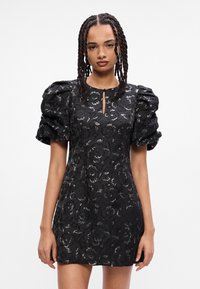 Crna haljina s napuhanim rukavima, cvjetnim uzorkom i detaljem u obliku ključa na vratu. Kratka dužina s sjajnim naglascima kroz tkaninu.