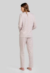 Mujer de pie descalza, de espaldas, vestida con una blusa de manga larga rosa claro y pantalones con un pequeño patrón de puntos rojos sobre fondo gris.