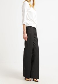 Pantaloni neri a gamba larga con una texture liscia, caratterizzati da una vita alta e sottili pieghe frontali, abbinati a una maglietta bianca a maniche lunghe.