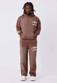 Felpa marrone con cappuccio e pantaloni della tuta abbinati in morbido tessuto, con accenti grafici di aerei di colore chiaro. Il modello è in piedi con le mani in tasca.