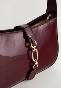 Burgundy patenteret lædertaske med en guldtonet kædeaccent, glat tekstur, lynlåslukning og en kontraststingdetalje på stroppen.