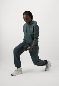 Tamsiai žalias hoody ir sportinės kelnės, pagamintos iš minkštos medžiagos su logotipu. Asmuo laiko juodą hantelį, dėvi šviesiai pilkus sportinius batelius.