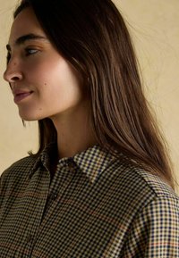 Pārbaudītas pogājamās krekls zaļā, zilā un bēšā krāsā ar klasisku apkakli. Audums izskatās teksturēts ar redzamām šuvēm un pogām.