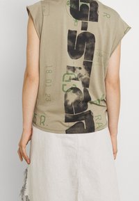 Person wearing a sleeveless beige shirt with bold black and green graphic text on the back and a white skirt with frayed edges.