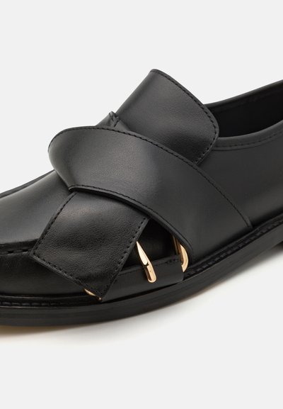 Μαύρο δερμάτινο παπούτσι slip-on με φαρδιά λωρίδα που διαθέτει χρυσά αξεσουάρ, λεία υφή και στρογγυλή μύτη.