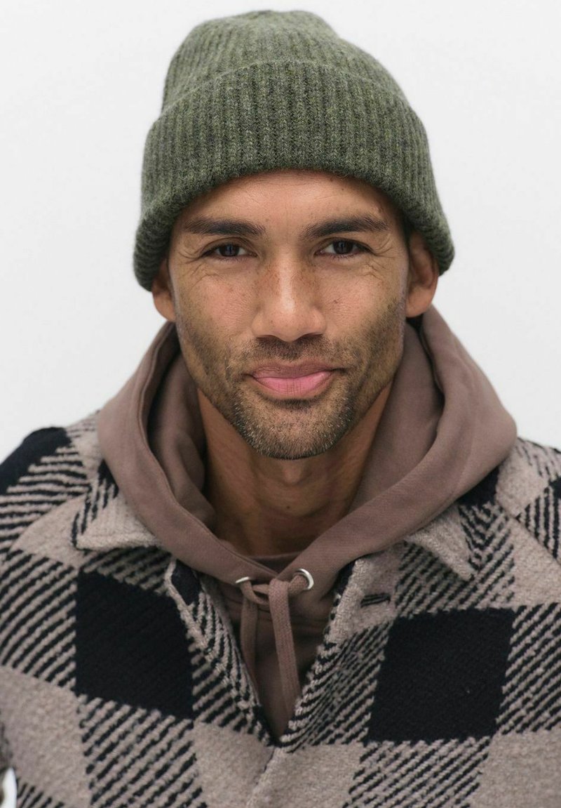Mand iført en grøn strikket beanie og en brun hoodie under en mønstret uldfrakke med sort og cremefarvede sektioner. Nærbillede portræt.