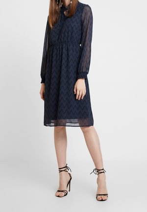 Woman wearing knee-length navy zigzag patterned dress with sheer long sleeves and black strappy high-heeled sandals.
