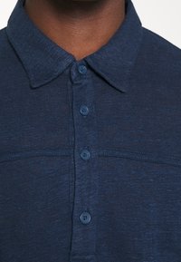 Námořnická modrá košile s límečkem vyrobená z texturované látky, s légou se pěti knoflíky a jemným pleteným vzorem.