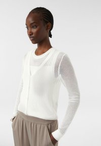 Weißer, gestrickter Cardigan mit V-Ausschnitt und Knopfverschluss, ausgestattet mit langen Ärmeln und gerippten Bündchen. Über einer hellen Unterlage und beigen Hosen getragen.