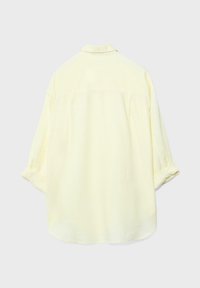 Jasnożółta koszula z długim rękawem i kołnierzykiem, pokazana od tyłu, rękawy luźno podwinięte przy mankietach, na jednolitym jasnym tle.