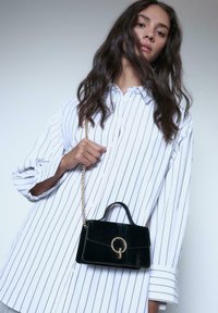 Borsa in velluto nero con manico superiore e tracolla a catena, caratterizzata da una chiusura circolare in oro, abbinata a una camicia bianca a righe.