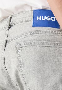 Jean en denim gris clair avec un patch en cuir bleu marqué "HUGO" sur la ceinture arrière, porté avec un haut blanc.