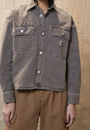 Person, die eine graue Knopfleiste-Jacke mit zwei vorderen Klappentaschen und braune Cargohosen vor einem strukturierten beigen Hintergrund trägt.
