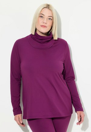 Blonde Frau, die ein lila langärmliges Rollkragenoberteil und passende Hosen trägt, steht vor einem weißen Hintergrund.