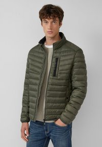 Leichte olivgrüne Steppjacke mit horizontaler Steppstruktur, Stehkragen und Reißverschlusstasche auf der Brust, kombiniert mit einem beigen Pullover.