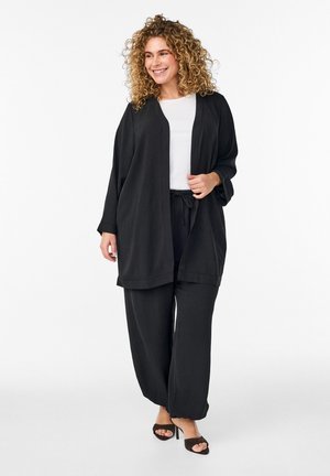 Usmievavá žena s kučeravými vlasmi, oblečená v čiernych širokých nohaviciach, voľnom čiernom kardigáne, bielom topán a čiernych otvorených sandáloch na vysokom opätku, na bielom pozadí.