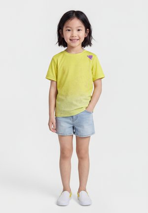 Hymyilevä lapsi, jolla on lyhyet mustat hiukset, ja joka pukeutunut keltaiseen t-paitaan, jossa on violetti kolmion muotoinen kuvio, vaaleisiin farkkushortseihin ja valkoisiin slip-on-kenkiin.