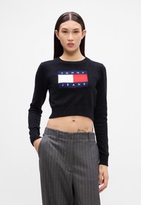 Černý zkrácený pletený svetr s žebrovanými manžetami, s obdélníkovým logem v modré, bílé a červené barvě s nápisem „TOMMY JEANS“. 