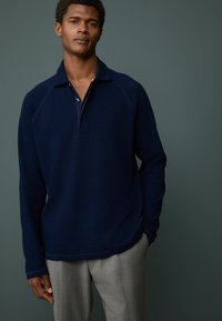 Marineblå pullover med et tekstureret mønster, der har krave, knaplukning og raglanærmer, båret med lys grå bukser.