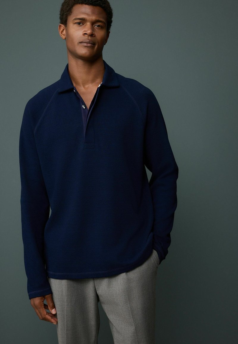 Marineblauwe pullover met een ribbelstructuur, een kraag en een verborgen knoopsluiting. Gecombineerd met grijze broeken, met een aansluitend design en lange mouwen.