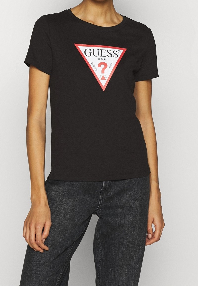 Personne portant un t-shirt noir Guess avec un logo triangle rouge et blanc et un jean noir en denim sur un fond uni.