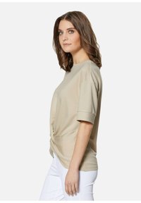 Beige top met korte mouwen en een ronde halslijn, met een gedraaide knoopdetail aan de voorkant, gemaakt van zachte, gladde stof, gecombineerd met witte broek.