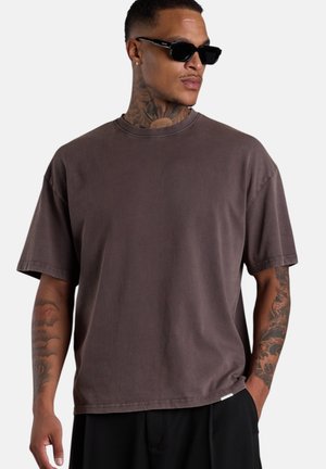 Mann mit Tattoos trägt ein dunkelbraunes oversized T-Shirt, schwarze Hose und schwarze Sonnenbrille, posiert mit einer Hand in der Tasche vor einfarbigem Hintergrund.