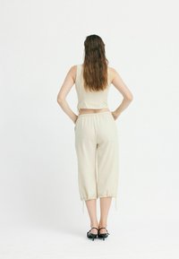 Beige, kurzes Tanktop mit verstellbarem Band, kombiniert mit leichten, taillierten Hosen mit Kordelzügen am Saum und schwarzen, hohen Sandalen.