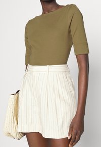 Oliivinvihreä, istuva paita lyhyillä hihoilla, yhdistetty valkoisiin, raidallisiin pleatattuihin shortseihin ja kudottuun beigeen käsilaukkuun.