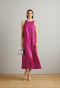 Fuchsiová maxi šaty s halter výstrihom z hladkého materiálu, s voľným strihom a bočnými vreckami. Strieborná kabelka pridáva kontrastný detail. Pozadie z drevenej podlahy.
